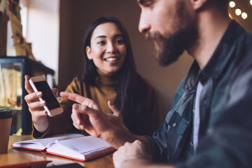 Cheerful Caucasian woman with cute smile on face showing social publication on smartphone to successful male colleague satisfied with content ideas, happy man and woman browsing mobile website