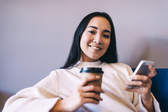 Positive young lady using smartphone while drinking hot coffee on couch