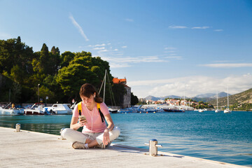 Smiling young pretty woman sitting in lotus pose on berth holding mobile phone with fantastic croatian sea coast view at background. Bright sunny day in Dubrovnik, Croatia. Travel lifestyle concept