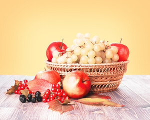 Fresh ripe fruits in a wicker basket, autumn leaves and berries on a wooden table on yellow gradient background, front view, copy space. Autumn harvest, Thanksgiving day concept. World vegan day.