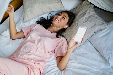 Happy Asian woman in pink sleepwear holding mockup smartphone technology laughing while listening positive music song during Sunday morning in home, joyful female in earphones using blank mobile phone