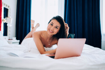 Obraz na płótnie Canvas Portrait of happy Asian blogger with modern cellphone and laptop technology smiling at camera during leisure time in hotel bedroom, cheerful freelancer with digital smartphone and netbook resting