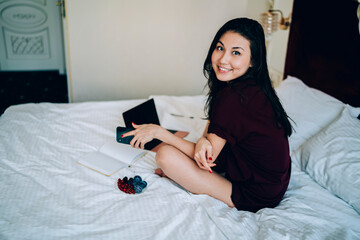 Portrait of happy Asian freelancer with laptop and smartphone devices smiling at camera during remote working in hotel bedroom, young hipster girl with mobile phone resting in home apartment