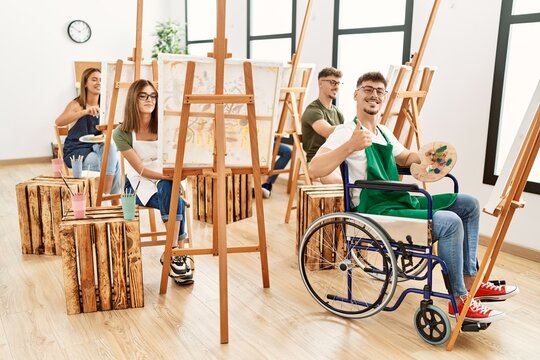 Young disabled man sitting on wheelchair drawing at art studio smiling happy and positive, thumb up doing excellent and approval sign