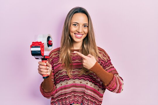 Beautiful hispanic woman holding packing tape smiling happy pointing with hand and finger