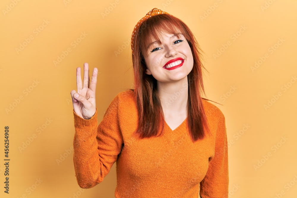 Wall mural Redhead young woman wearing casual orange sweater showing and pointing up with fingers number three while smiling confident and happy.