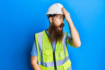 Redhead man with long beard wearing safety helmet and reflective jacket surprised with hand on head...