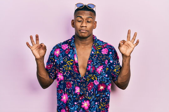 Young black man wearing hawaiian shirt and sunglasses relax and smiling with eyes closed doing meditation gesture with fingers. yoga concept.