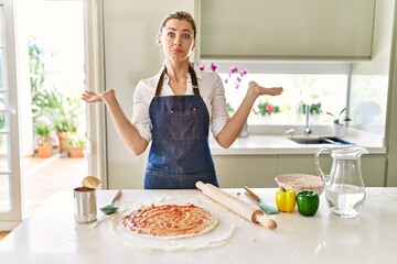 Beautiful blonde woman wearing apron cooking pizza clueless and confused expression with arms and...