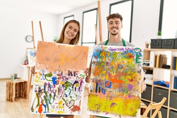 Young hispanic artist couple smiling happy showing canvas draw at art studio.