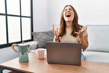 Young brunette woman using laptop at home drinking a cup of coffee crazy and mad shouting and yelling with aggressive expression and arms raised. frustration concept.