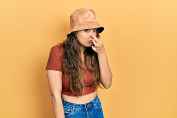 Young hispanic girl wearing casual clothes and hat smelling something stinky and disgusting, intolerable smell, holding breath with fingers on nose. bad smell