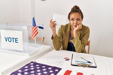 Beautiful hispanic woman holding voting envelop in ballot box serious face thinking about question...