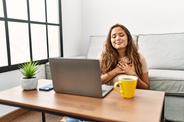 Beautiful hispanic woman using computer laptop at home smiling with hands on chest with closed eyes and grateful gesture on face. health concept.