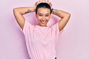 Beautiful hispanic woman with short hair wearing casual pink t shirt posing funny and crazy with fingers on head as bunny ears, smiling cheerful