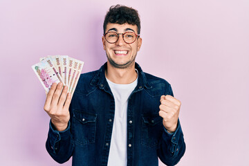 Young hispanic man holding saudi arabia riyal banknotes screaming proud, celebrating victory and success very excited with raised arm