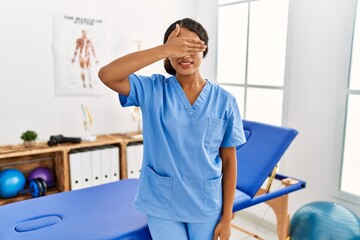 Beautiful hispanic physiotherapist woman at pain recovery clinic smiling and laughing with hand on face covering eyes for surprise. blind concept.