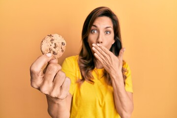 Young latin woman holding cookie covering mouth with hand, shocked and afraid for mistake....