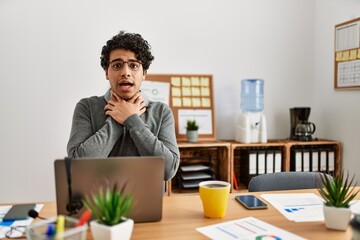 Young hispanic man wearing business style sitting on desk at office shouting and suffocate because painful strangle. health problem. asphyxiate and suicide concept.