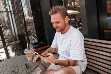 Obraz na płótnie Canvas Handsome young blonde man sitting on cafe terrace with coffee beverage smiling with mobile smartphone on big city street glass building reflection background. Millennial hipster on summer sunny day