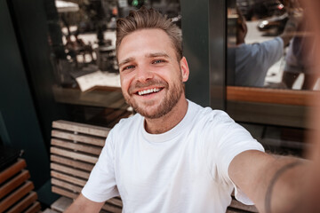 Handsome young blonde making smiling selfie wears white t-shirt in cafe terrace table with coffee beverage holding mobile smartphone. Close up male portrait. Hipster stylish millennial with hairstyle