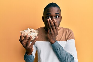 Young african american man holding bowl with pumpkin seeds covering mouth with hand, shocked and...