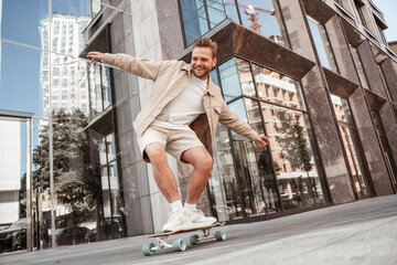 Handsome blonde young man doing tricks on skateboard trains on longboard outdoors at street with urban background. Excited laughing sporty skater practices outside. Extreme sport lifestyle concept. © Marharyta Hanhalo