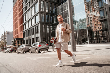 Attractive man skateboarder holding longboard on shoulder and cup of coffee takeaway looks seriously focused at distance while crossing the road on urban building background. Wears stylish outfit.