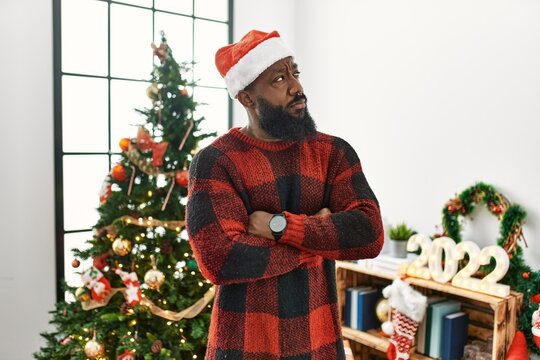 African american man wearing santa claus hat standing by christmas tree looking to the side with arms crossed convinced and confident