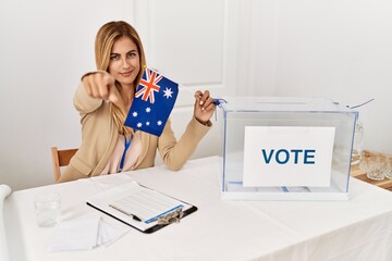 Blonde beautiful young woman at political campaign holding australian flag pointing with finger to the camera and to you, confident gesture looking serious