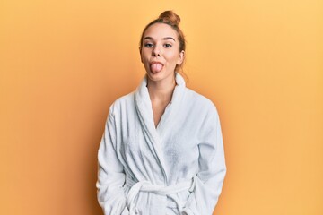 Young blonde woman wearing bathrobe sticking tongue out happy with funny expression. emotion...