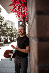 Attractive blonde young man standing against brick city wall holding wooden longboard smiling laughing while using smartphone, posting to social media on city landscape background. Leisure concept.