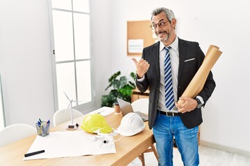 Middle age hispanic business man holding paper blueprints pointing to the back behind with hand and thumbs up, smiling confident