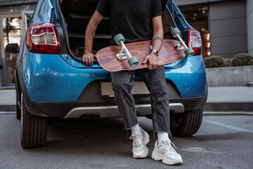 Faceless portrait of young stylish man leaning on blue car trunk holding longboard wearing sneakers. City life concept. Extreme sports concept, outdoors leisure concept. Hipster concept. Skateboarder.