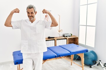 Obraz premium Middle age hispanic therapist man working at pain recovery clinic showing arms muscles smiling proud. fitness concept.
