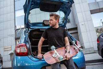 Attractive european man holding longboard sitting in modern car trunk, looks at distance, wearing...