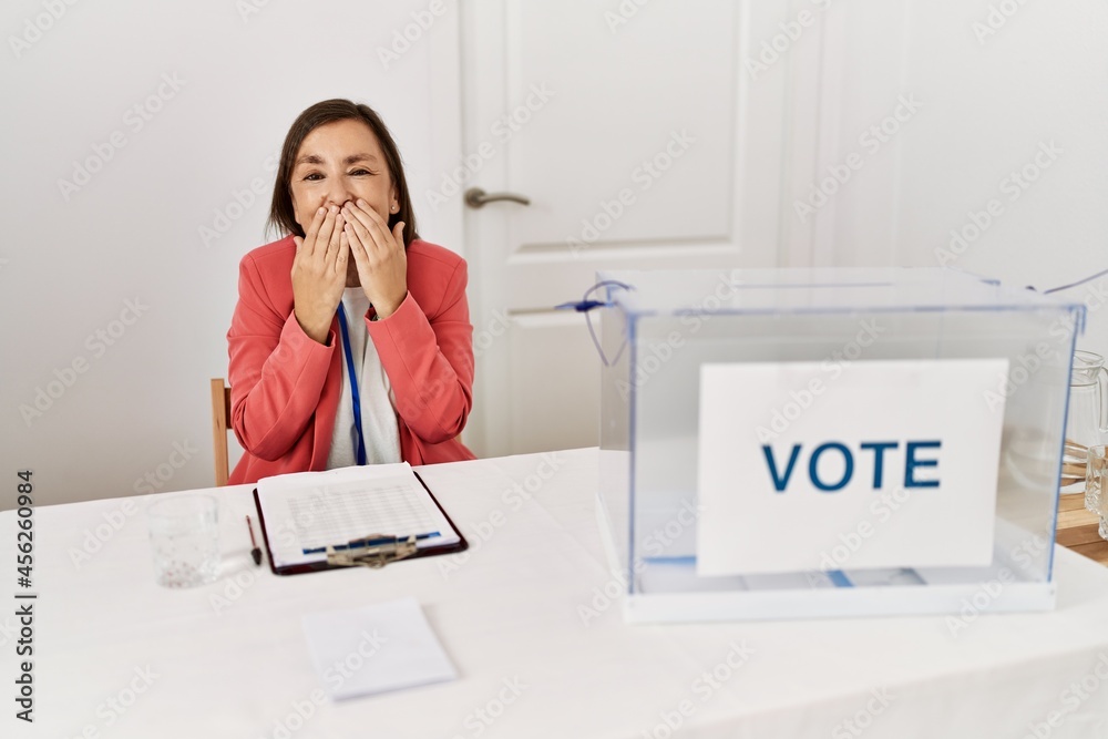 Wall mural Beautiful middle age hispanic woman at political election sitting by ballot laughing and embarrassed giggle covering mouth with hands, gossip and scandal concept