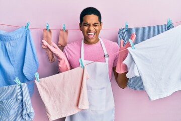 Young handsome hispanic man wearing cleaner apron holding clothes on clothesline celebrating mad...