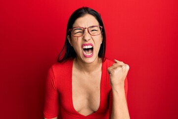 Young latin woman wearing casual clothes and glasses angry and mad raising fist frustrated and furious while shouting with anger. rage and aggressive concept.