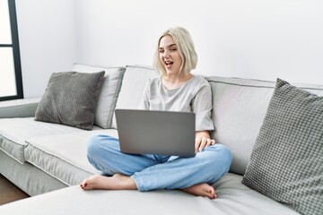 Young caucasian woman using laptop at home sitting on the sofa winking looking at the camera with...