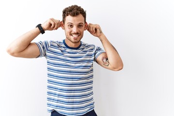 Handsome young man standing over isolated background smiling pulling ears with fingers, funny...