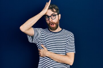 Caucasian man with beard wearing striped t shirt and glasses surprised with hand on head for mistake, remember error. forgot, bad memory concept.