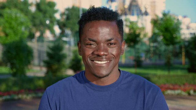 Close up smiling afro-american man looks camera on background beautiful park in summer. Portrait happy African male student wears blue t-shirt