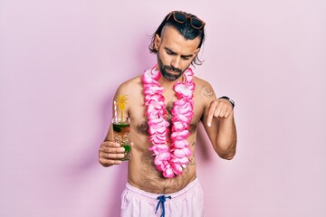 Young hispanic man wearing swimsuit and hawaiian lei drinking tropical cocktail pointing down...