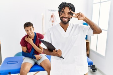 Young hispanic man working at pain recovery clinic with a man with broken arm doing peace symbol with fingers over face, smiling cheerful showing victory