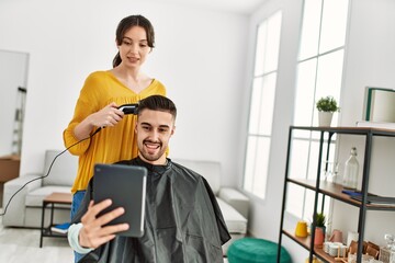 Fototapeta premium Young hispanic man looking touchapd screen while hairdresser woman cut hair at beauty center.