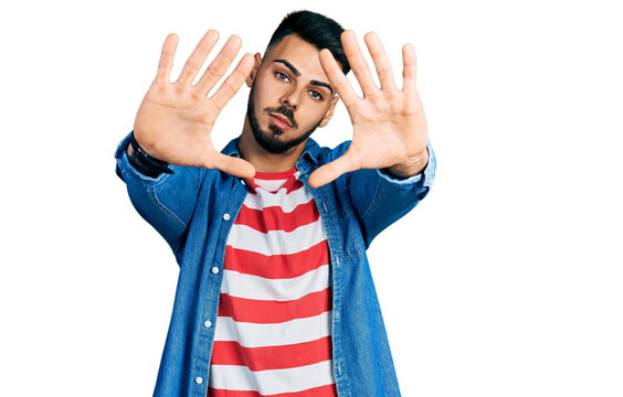 Young hispanic man with beard wearing casual denim jacket doing frame using hands palms and fingers, camera perspective