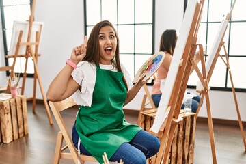 Young hispanic artist women painting on canvas at art studio smiling amazed and surprised and pointing up with fingers and raised arms.