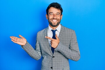 Handsome man with beard wearing business suit and tie amazed and smiling to the camera while presenting with hand and pointing with finger.