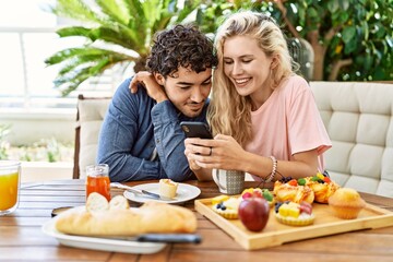 Young couple having breakfast and using smartphone sitting on the table at terrace.